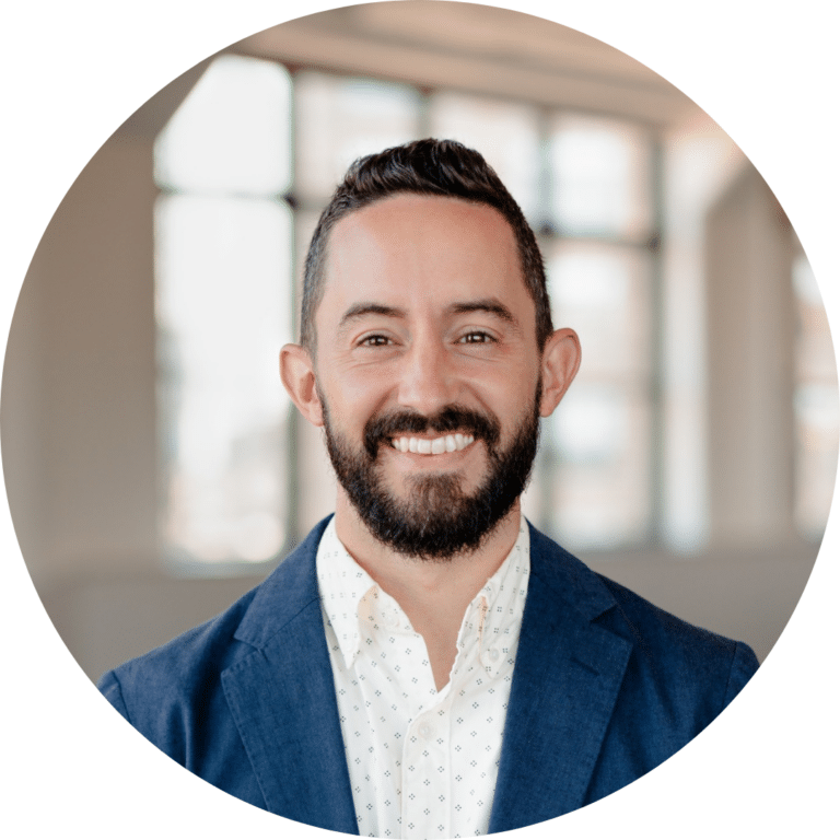 A man with a beard is smiling and wearing a blue blazer over a white shirt. He is indoors with large windows in the background.