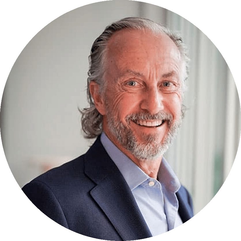 A man with gray hair and a beard, wearing a blue shirt and dark blazer, smiles while looking at the camera.